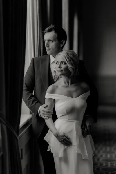 Black and white image of a bride and groom standing by a large sash window in a grand interior