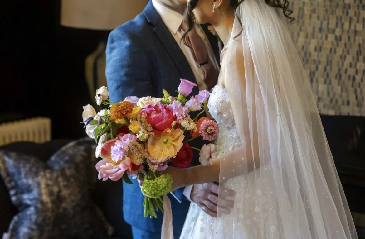 Close-up of a bride holding a vivid, abundant bouquet of hot pink peonies, orange poppies, marigolds, lilac tulips, red ranunculus, green viburnum and white scabiosa against a pearl-embellished wedding dress, groom in a blue suit standing behind her.