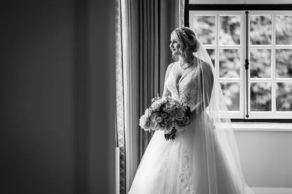 Black and white portrait of a bride in a lace ballgown and cathedral veil standing at a sash window holding a full, rounded bouquet of garden roses and ranunculus, looking out to the right.