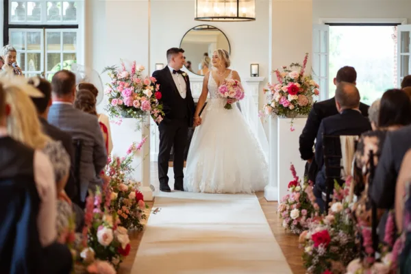 Bride and groom walking back down the aisle of a bright ceremony room lined with abundant floor-standing arrangements and aisle clusters of hot pink, coral and blush flowers including roses, peonies, foxglove and ranunculus, guests seated either side.