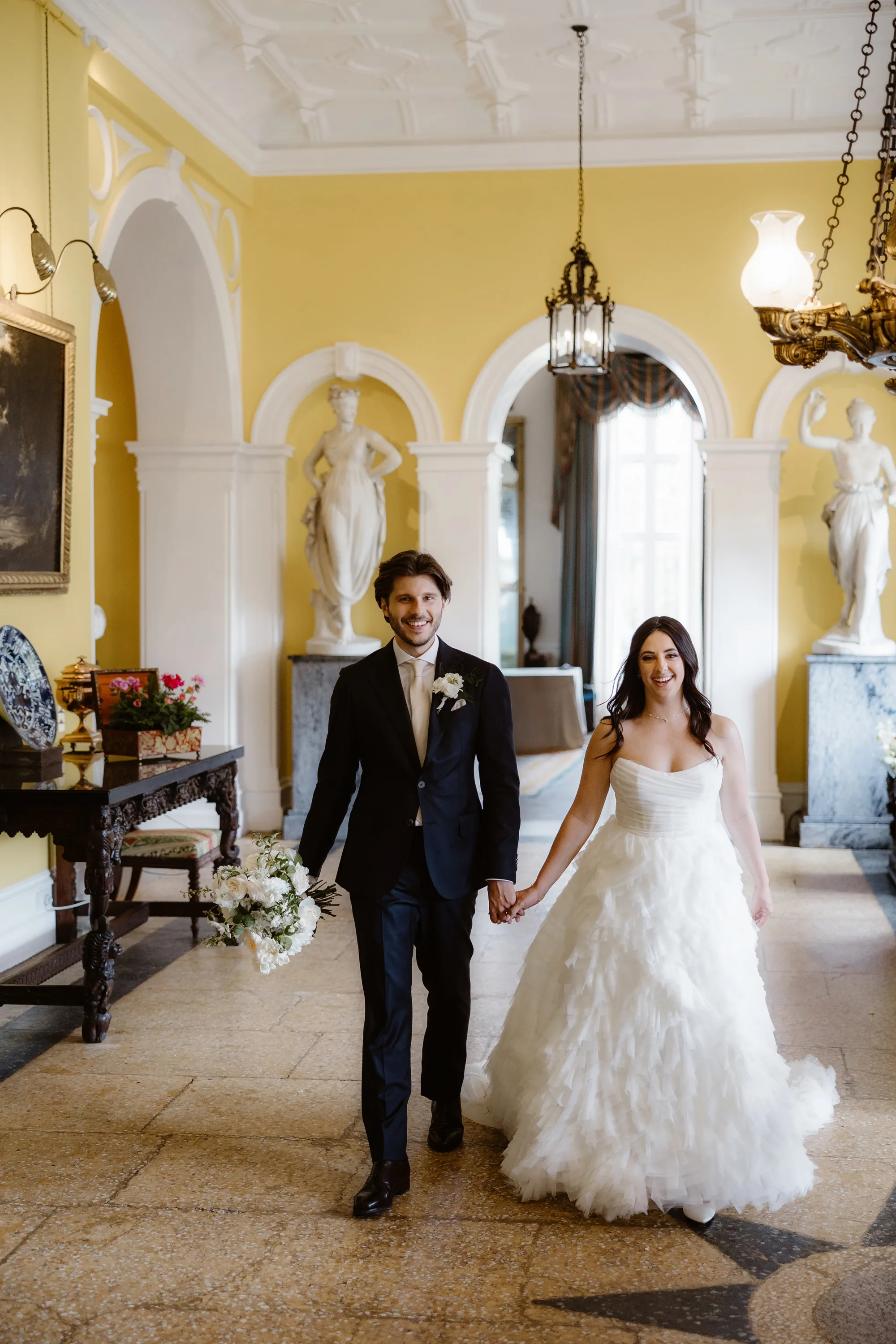 Sophie Dimitri Wedding Day Jono Symonds Photo Film 414 - Sophie & Dimitri's Beautiful, Relaxed & Elegant Wedding Weekend at Capesthorne Hall in Cheshire Elegant portrait of a bride and groom holding hands as they gently stroll through the interior of Capesthorne Hall in Cheshire on their wedding day. He wears a dark blue Moss Bros suit and carries the brides bouquet - the bride wears a frilly dress by Watters and has her hair long and loose.
