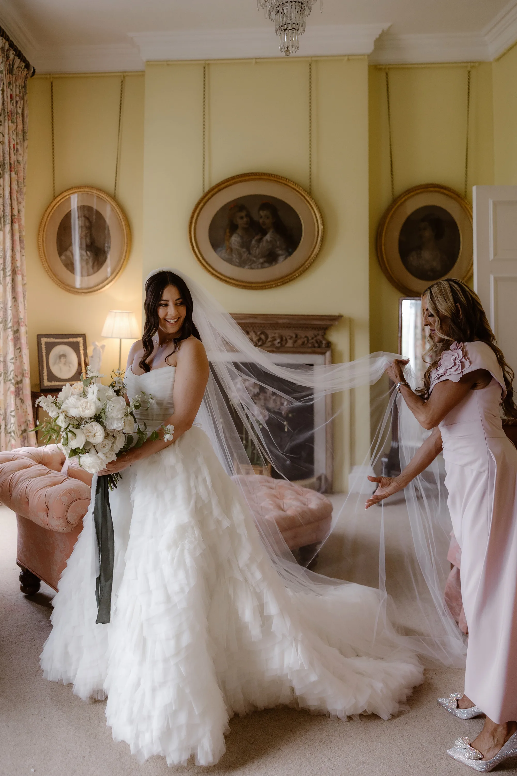 Sophie Dimitri Wedding Day Jono Symonds Photo Film 89 - Sophie & Dimitri's Beautiful, Relaxed & Elegant Wedding Weekend at Capesthorne Hall in Cheshire Bride being assisted with her veil on the morning of her wedding at Capesthorne Hall in Cheshire. She wears a Watters dress with frilly tier and carries a large white bouquet.
