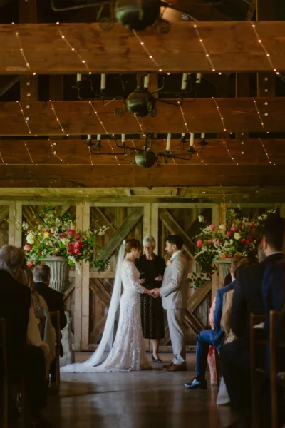 A ceremony in Dewsall Court's Wainhouse barn, with flowers at the altar that earned their place — two generous stone urn arrangements, packed with British-grown tulips, ranunculus, coral roses and white blossom in a palette that felt like the first proper day of spring. The rustic timber backdrop and fairy-lit ceiling did the rest. Floristry by Bloomify.