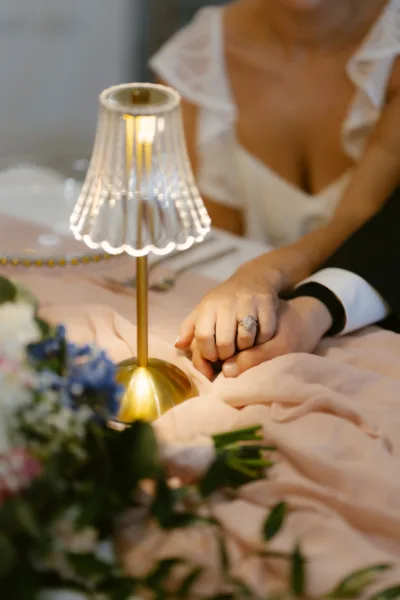 Close-up of hands reaching towards a glowing gold table lamp surrounded by blue wedding florals