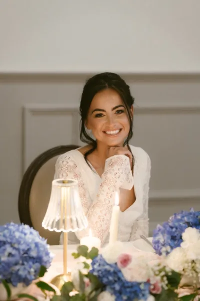 Smiling bride in a lace wedding dress seated at a candlelit reception table with blue hydrangeas