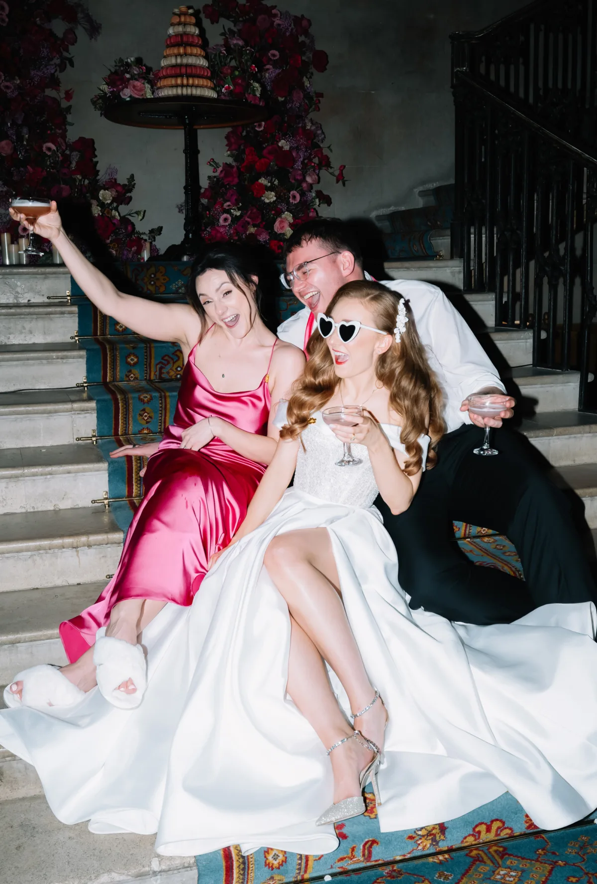 Bride celebrating with her bridesmaid and groom on the grand staircase at Ashridge House during the evening wedding reception.