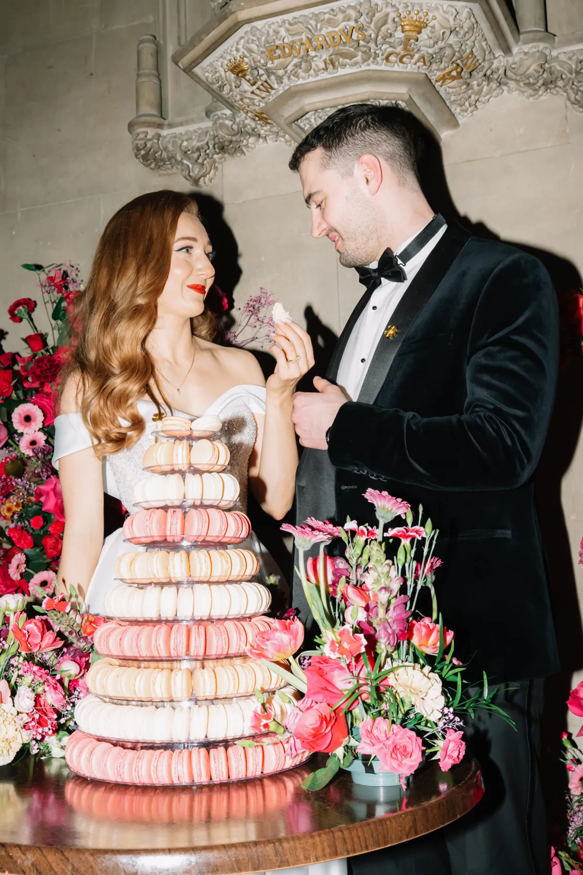 Bride and groom sharing macarons beside their macaron wedding cake, surrounded by vibrant pink floral arrangements at their Ashridge House wedding reception.