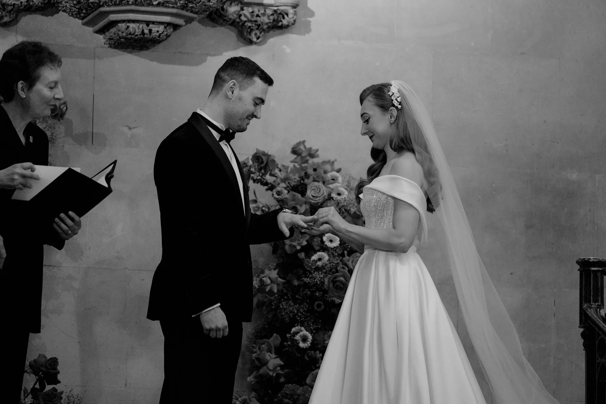 Bride placing a wedding ring on the groom's finger during their ceremony at Ashridge House in Hertfordshire.