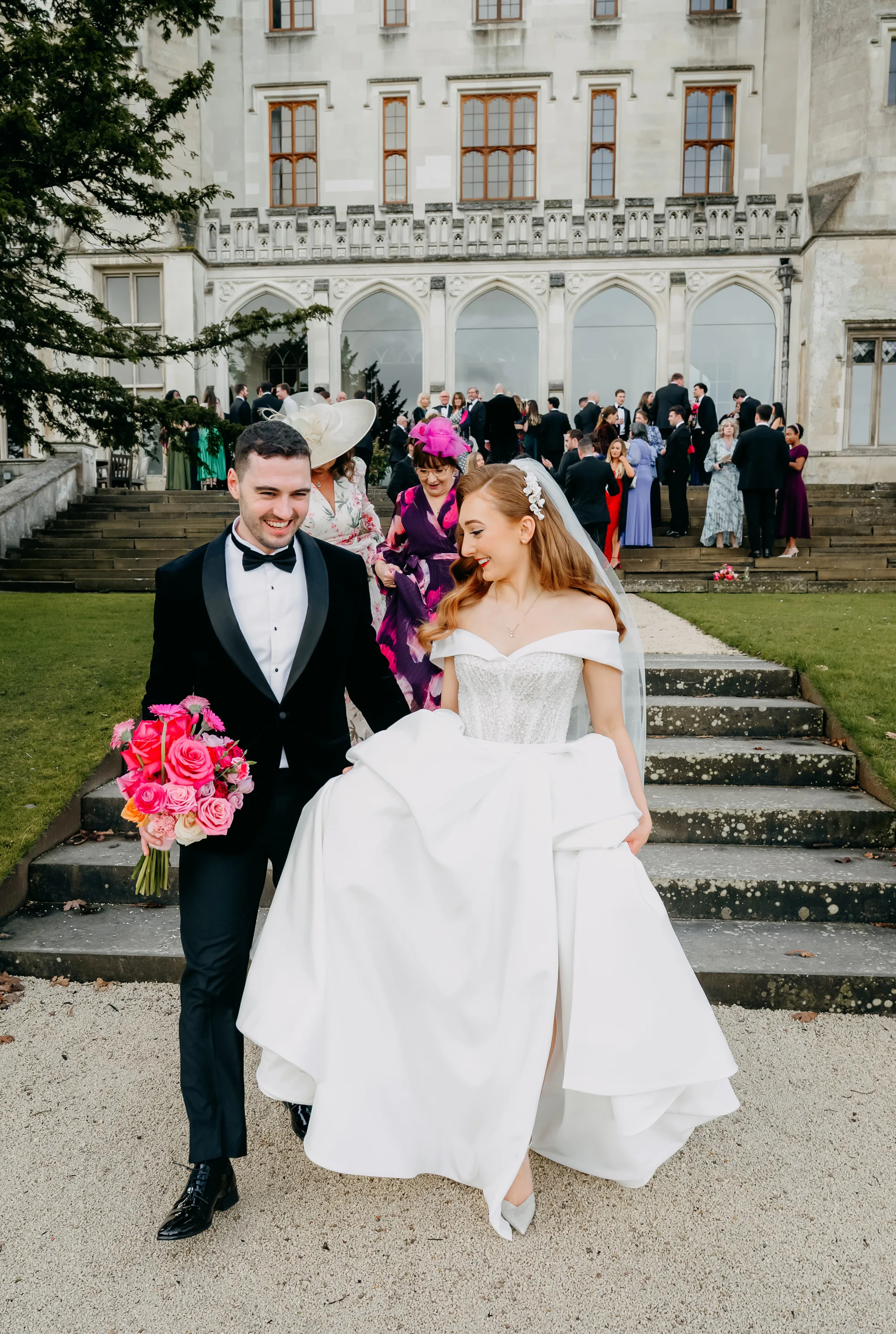 Bride and groom walking through the grounds of Ashridge House after their wedding ceremony, with guests celebrating behind them.
