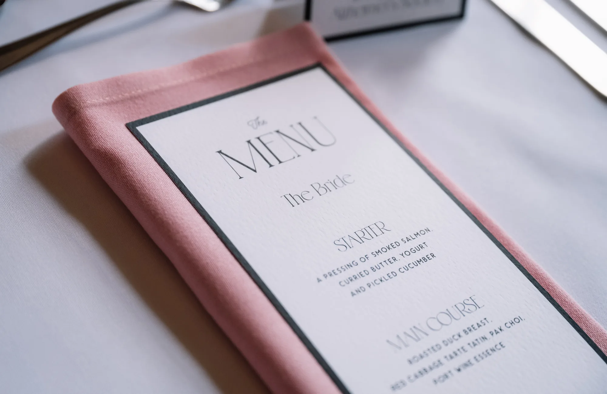 Wedding breakfast menu featuring Art Deco inspired font with blush pink linen napkin styling at a place setting for the reception at Ashridge House in Hertfordshire.