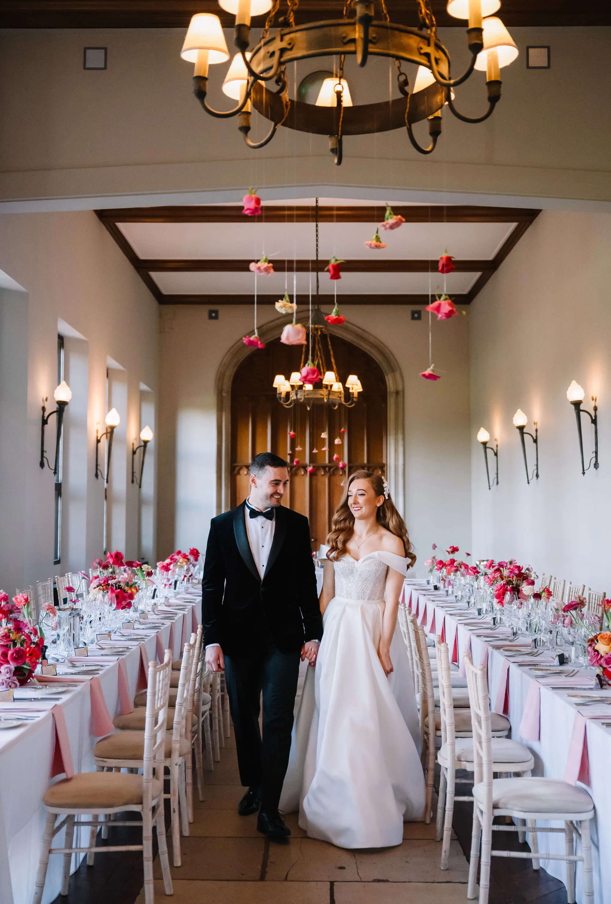 Bride and groom walking through their wedding breakfast room at Ashridge House with vibrant pink floral styling by Hanako Flowers, including flower heads hanging from the ceiling.