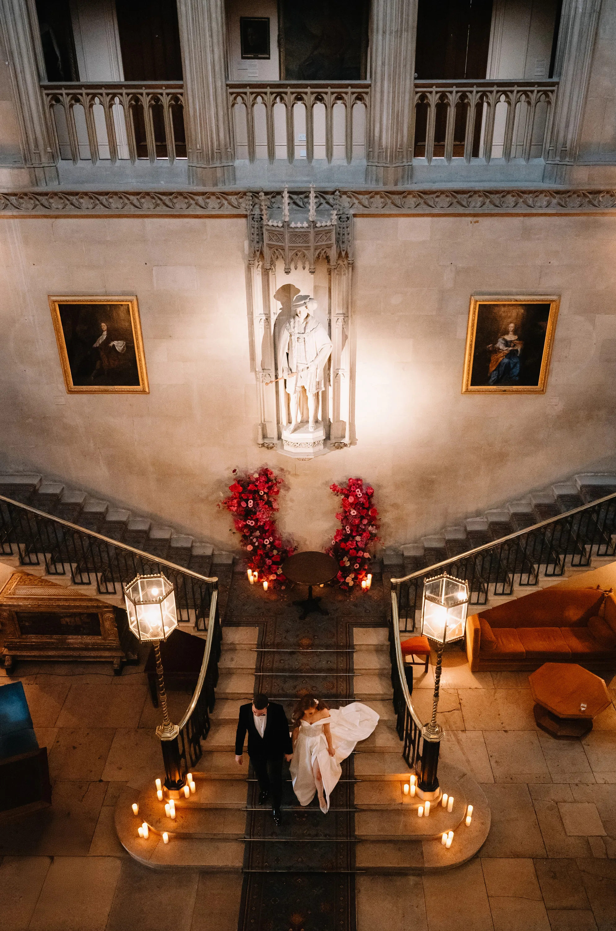 Overhead view of the bride and groom leaving their ceremony beneath the grand staircase at Ashridge House, surrounded by candlelight and vibrant pink floral displays.