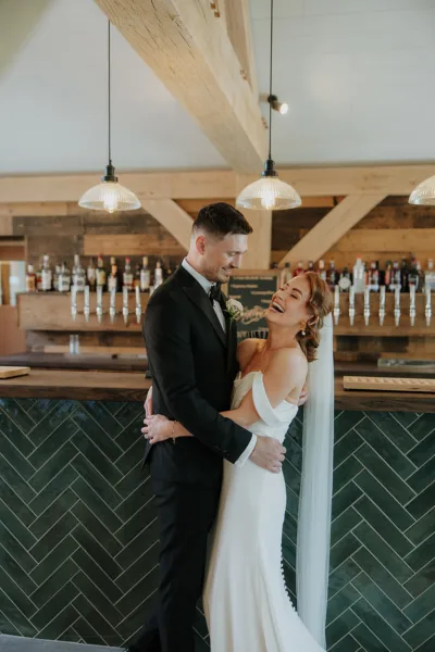 Newlyweds laugh in front of a stylish bar in a Kent wedding barn