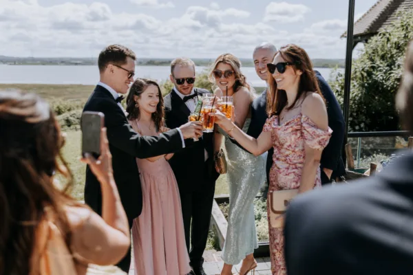 Wedding guests raise a glass during a waterside drinks reception at The Harty Estate wedding venue