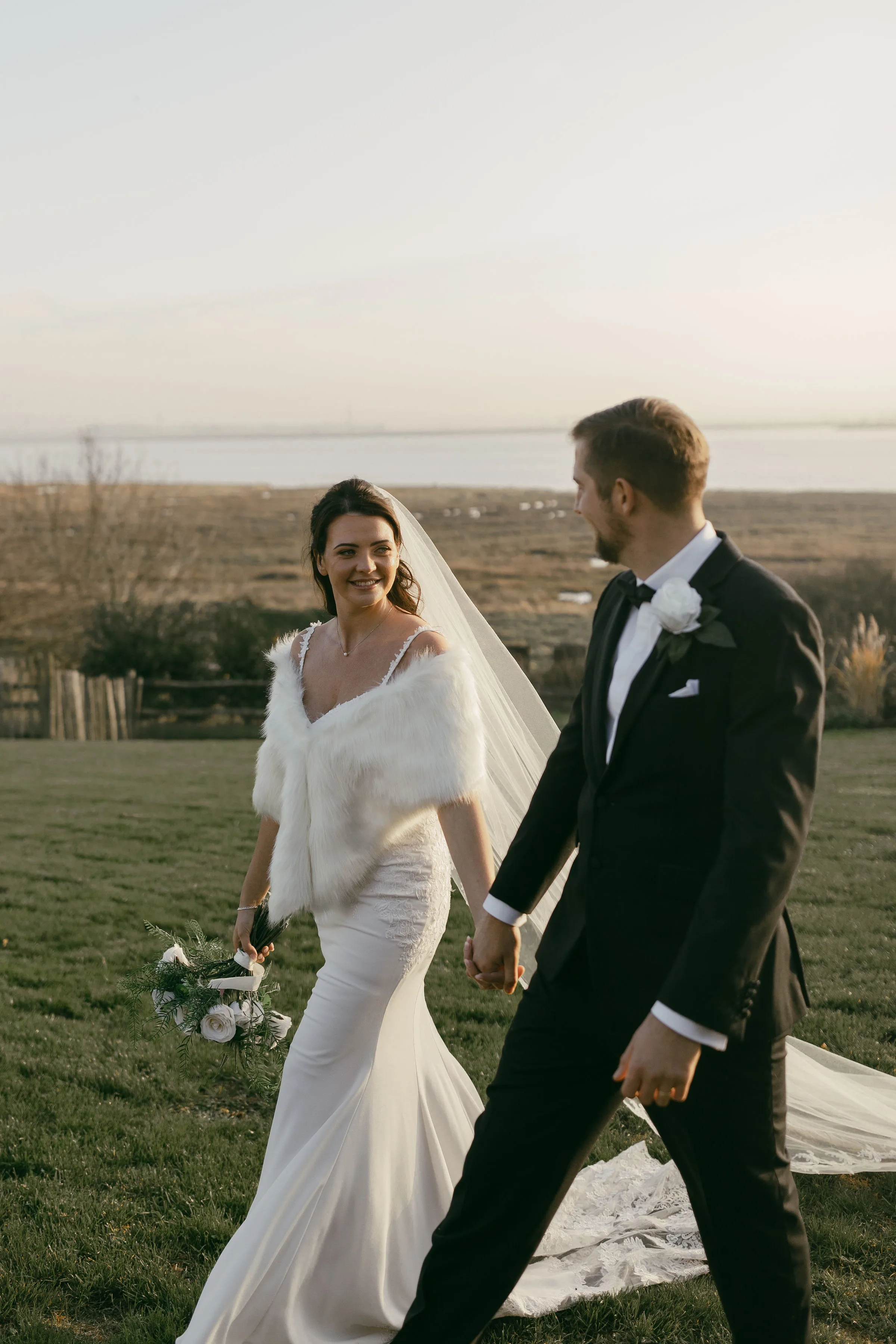Bride and groom walk hand in hand on their winter wedding day, with the Kent coast behind