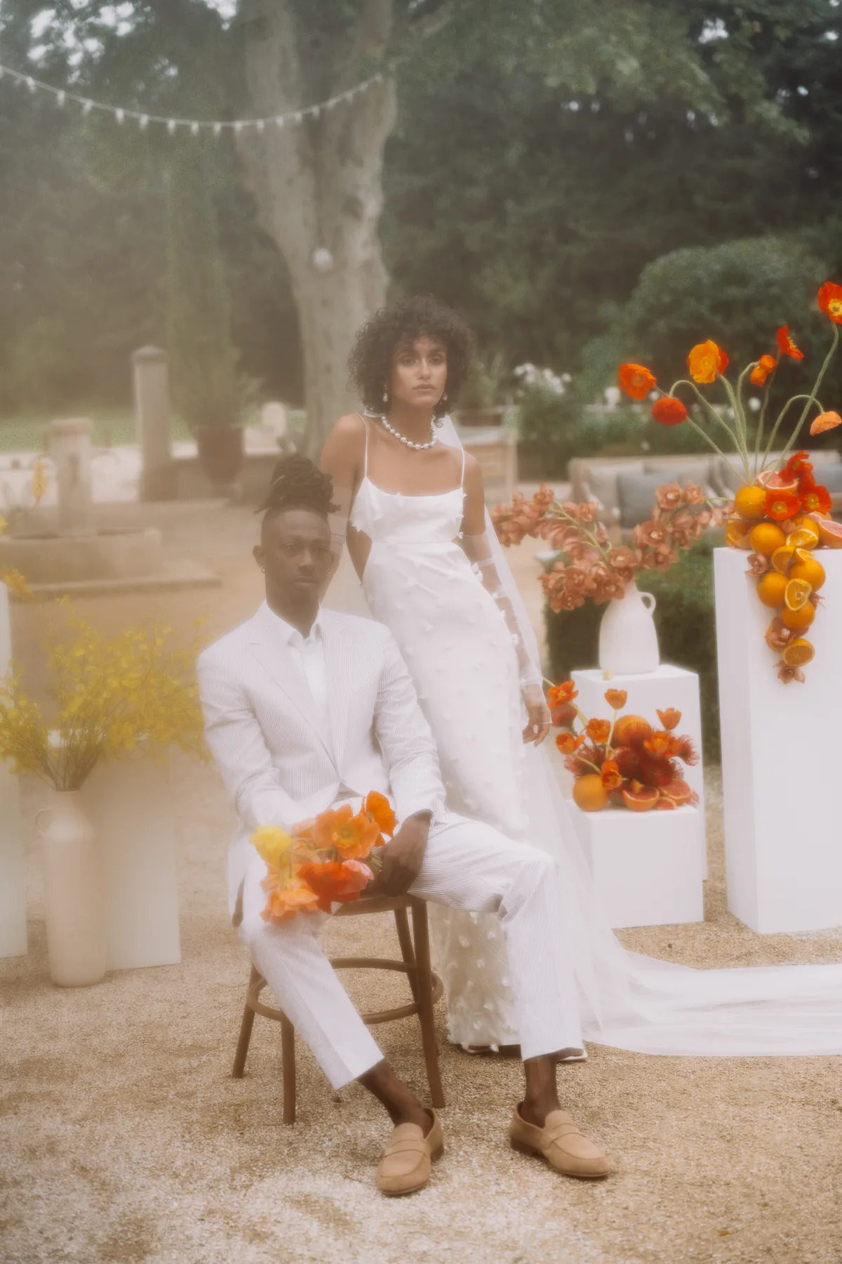 Model Black bride and groom seated in the grounds of Le Mas d'Arvieux in Provence, France, surrounded by white plinths displaying vibrant and coourful fruit and flowers like oranges and Icelandic Poppies.