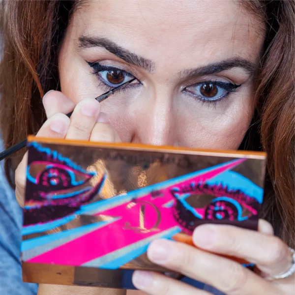 Close-up of a woman applying eyeliner while looking into a colourful mirror.
