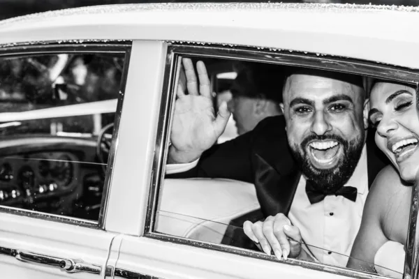 Laughing bride and groom waving from the window of their wedding car.