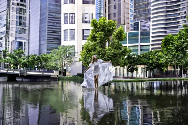 Bride in a flowing white dress standing beside water in a modern city setting.