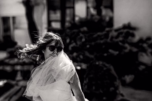 Black and white candid portrait of a bride in sunglasses with her veil blowing in the wind.
