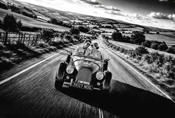 Bride and groom travelling through the countryside in a classic open-top sports car on their wedding day.