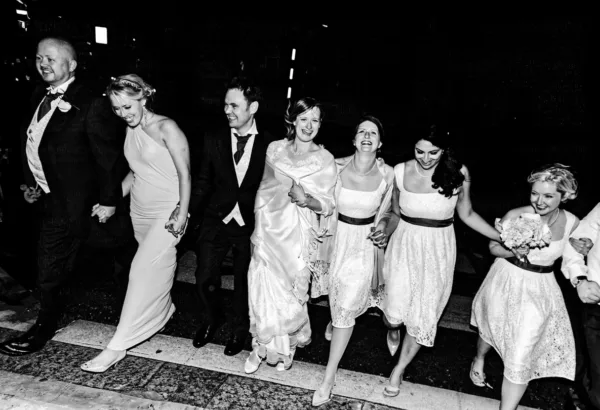 Bride, groom and wedding party laughing and walking together at night in a candid black-and-white photograph.