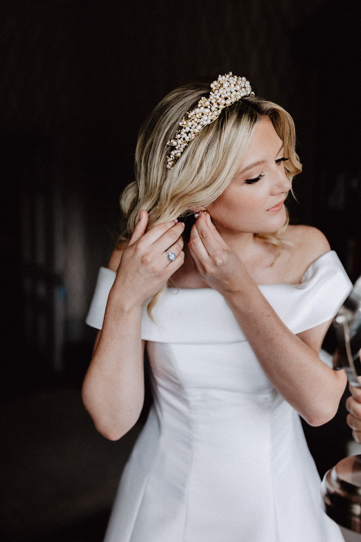 Bride wearing a pearl headband and an off-the-shoulder wedding dress, fastening an earring as she gets ready.