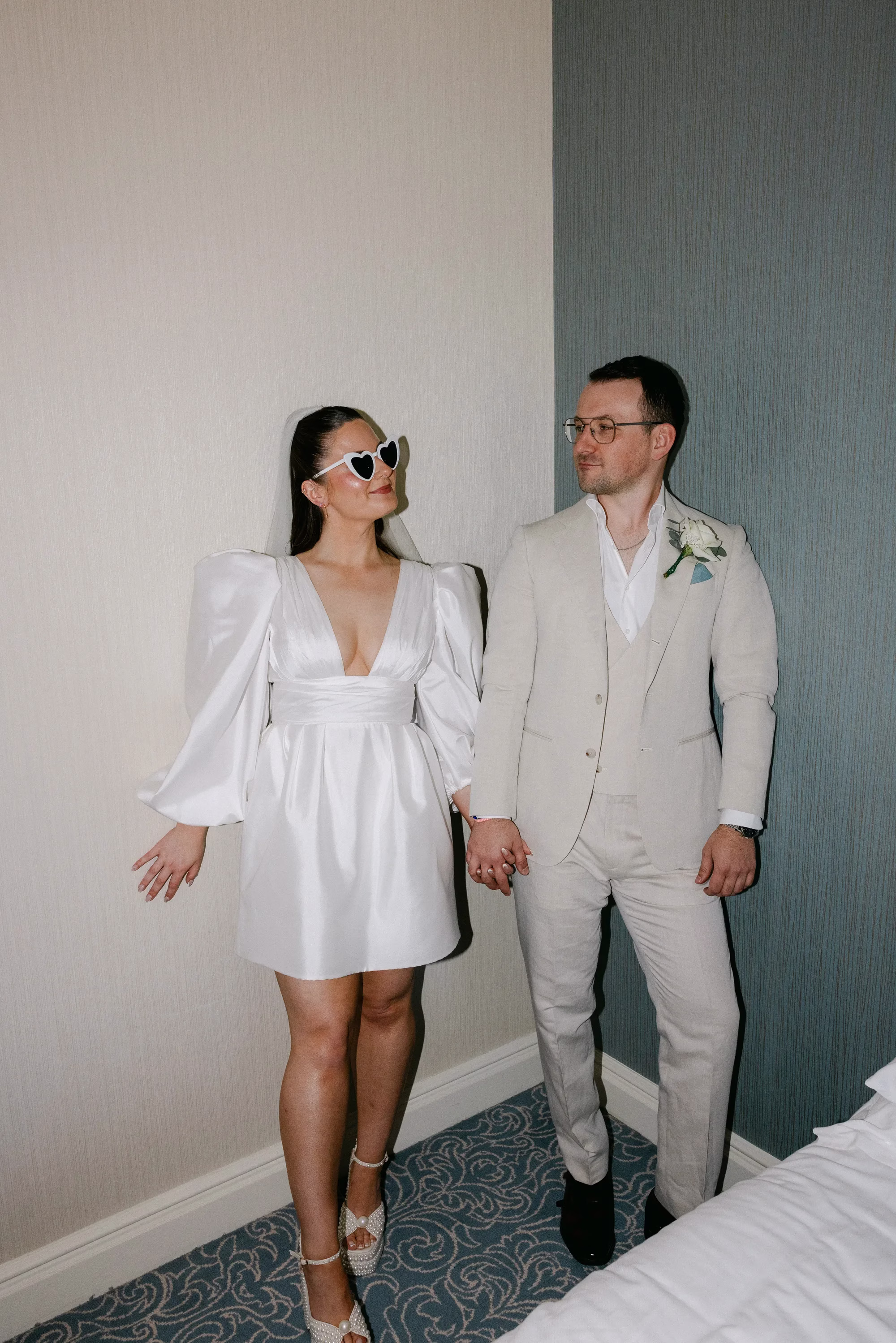 Bride in heart-shaped sunglasses and a short puff-sleeved wedding dress holding hands with the groom in a cream three-piece suit.