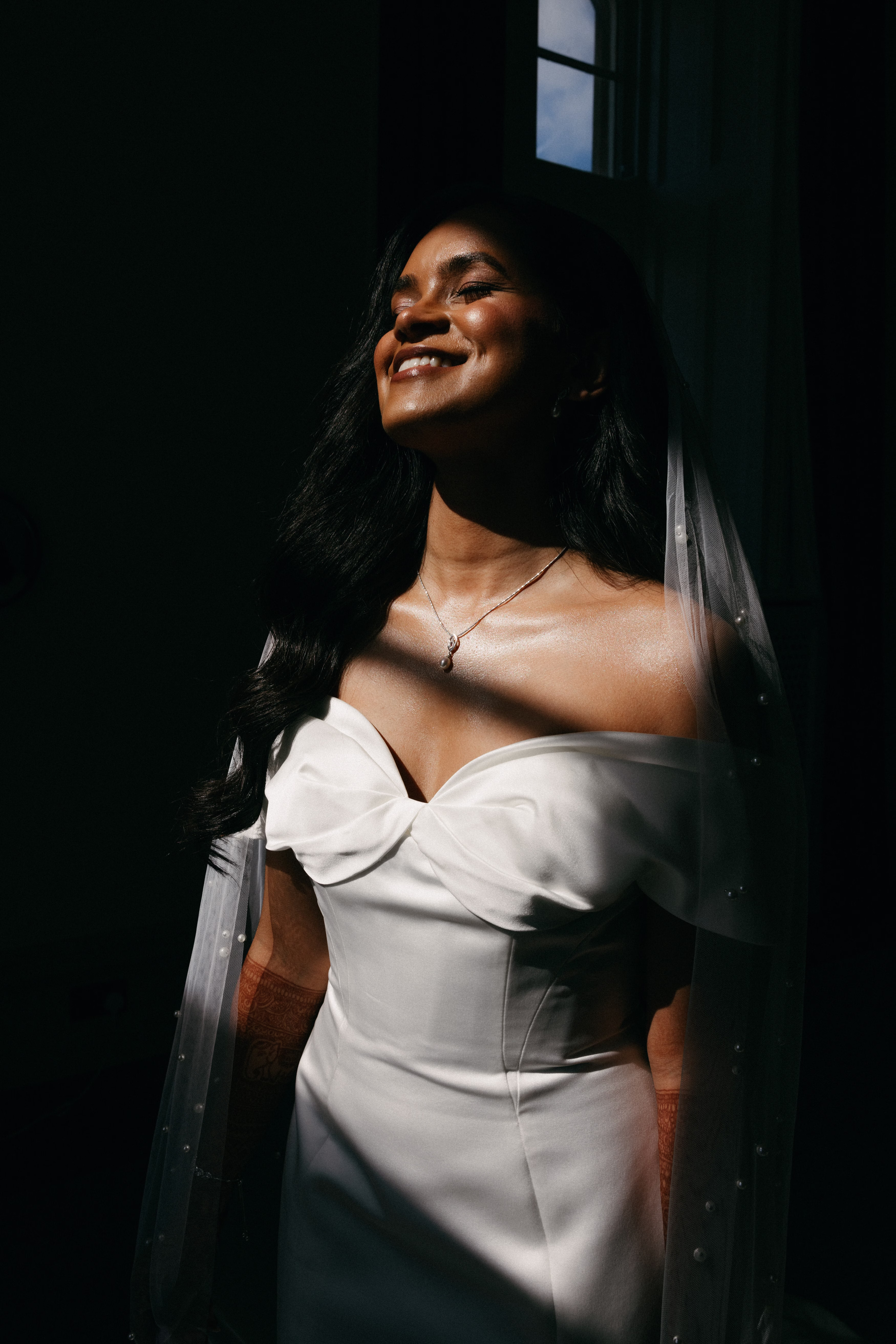 Bride smiling in a shaft of sunlight, wearing an off-the-shoulder wedding dress and a pearl-dotted veil against a dark background.
