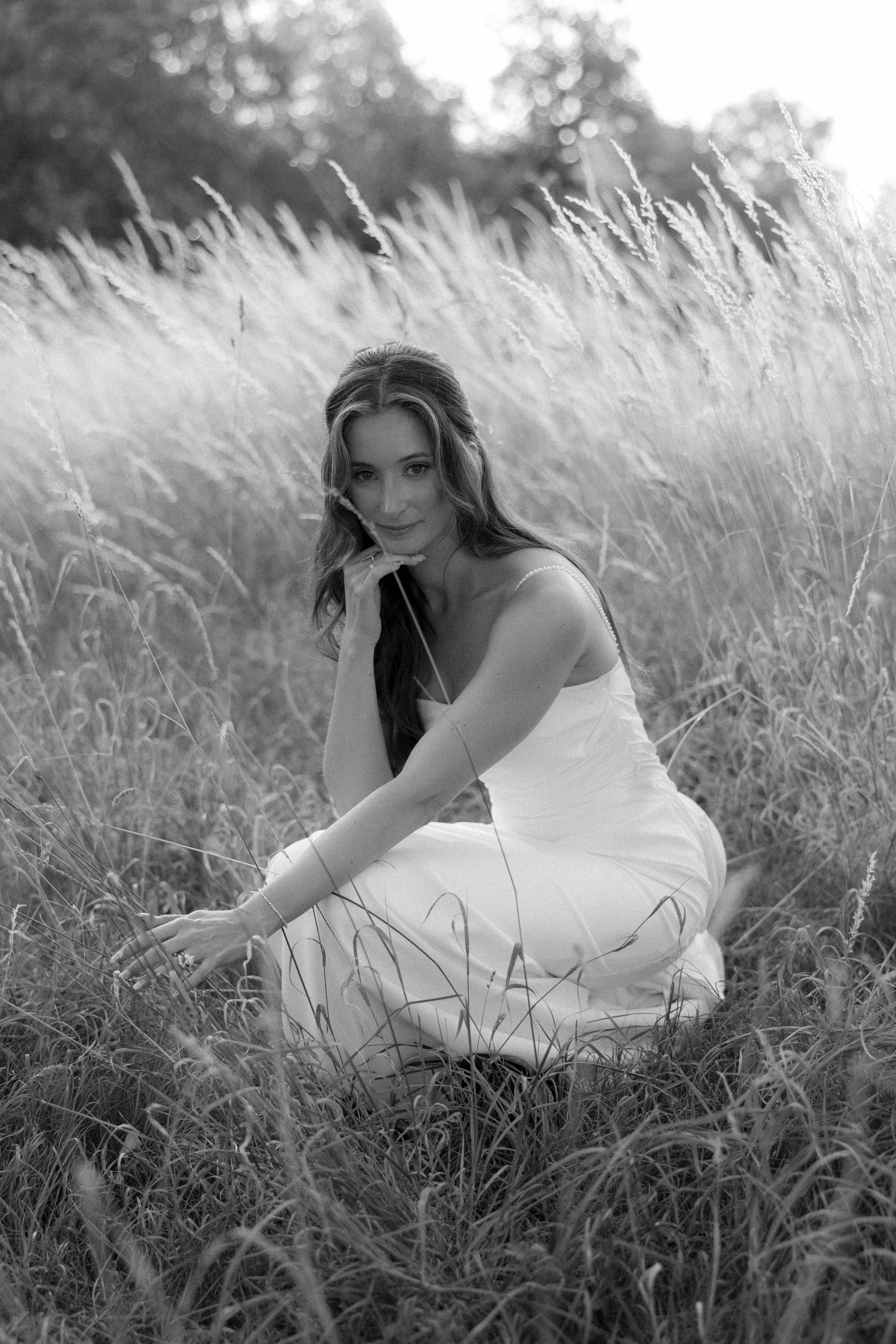 Black and white portrait of a bride in a thin-strapped wedding dress, seated among tall grasses with her chin resting on her hand.