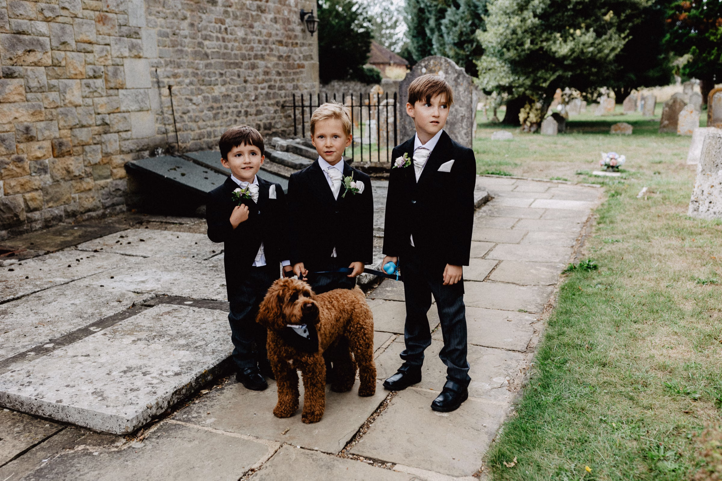 Three cute page boys in dark suits and pale cravats standing outside a stone church with a curly-haired brown dog on a lead.