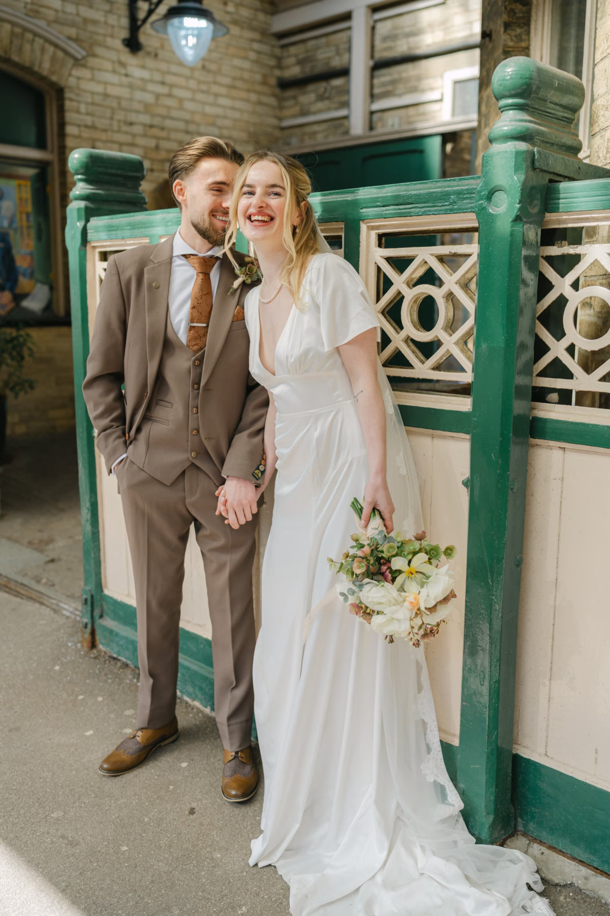 Bride in a flowing Wilderness Bride gown and groom in a brown suit holding hands and laughing beside green station railings