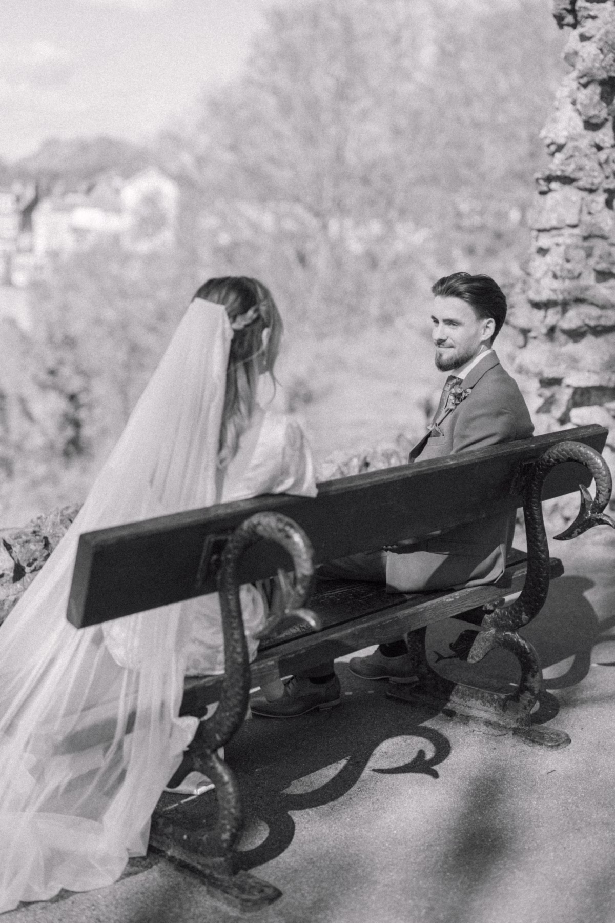 Black and white portrait of a couple sitting on a wooden bench, with the bride’s long veil draped over the back