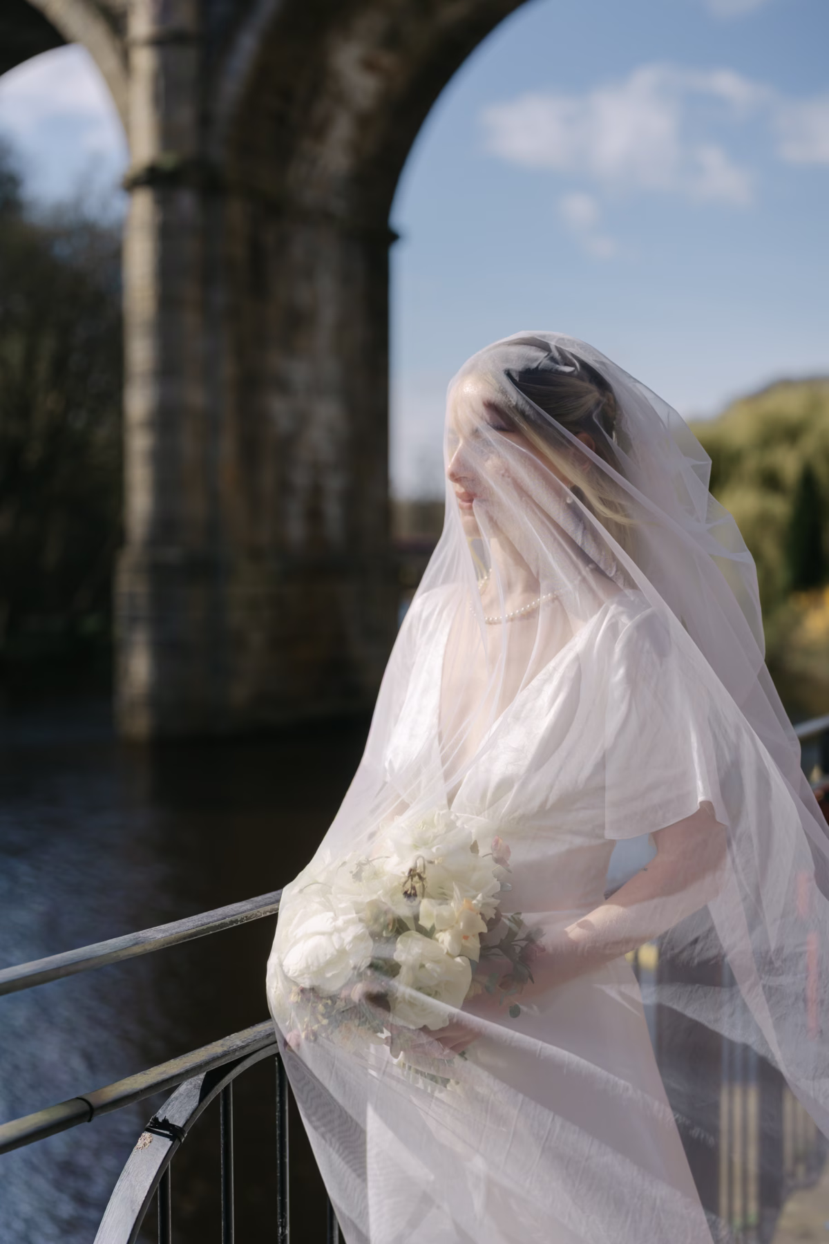 Bride beneath a sheer veil holding a white bouquet beside the river, with Knaresborough viaduct behind