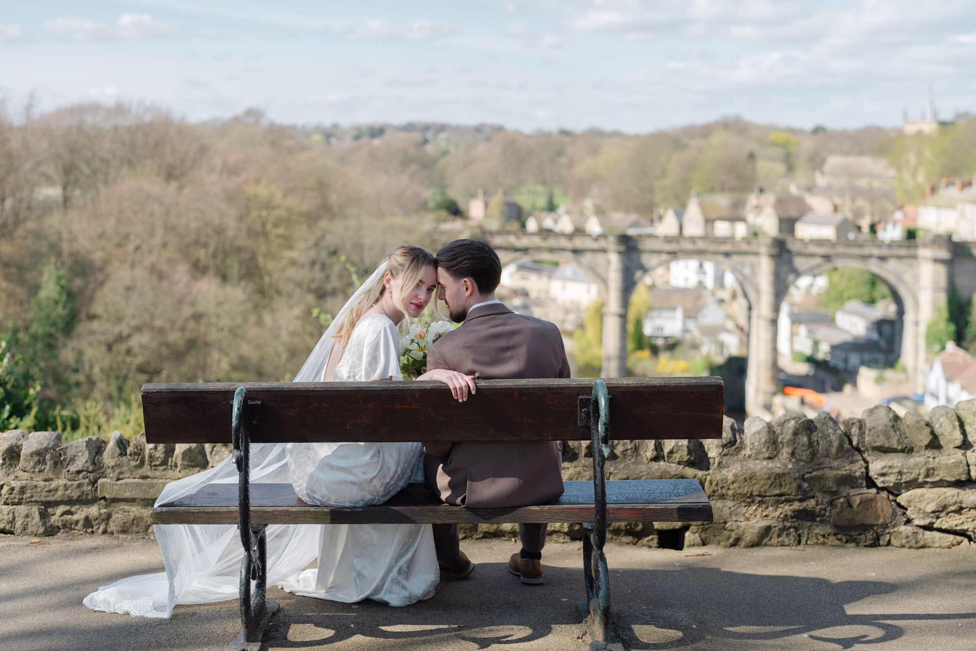 Groom touching the smiling bride’s cheek as they sit together on a wooden bench with her spring bouquet