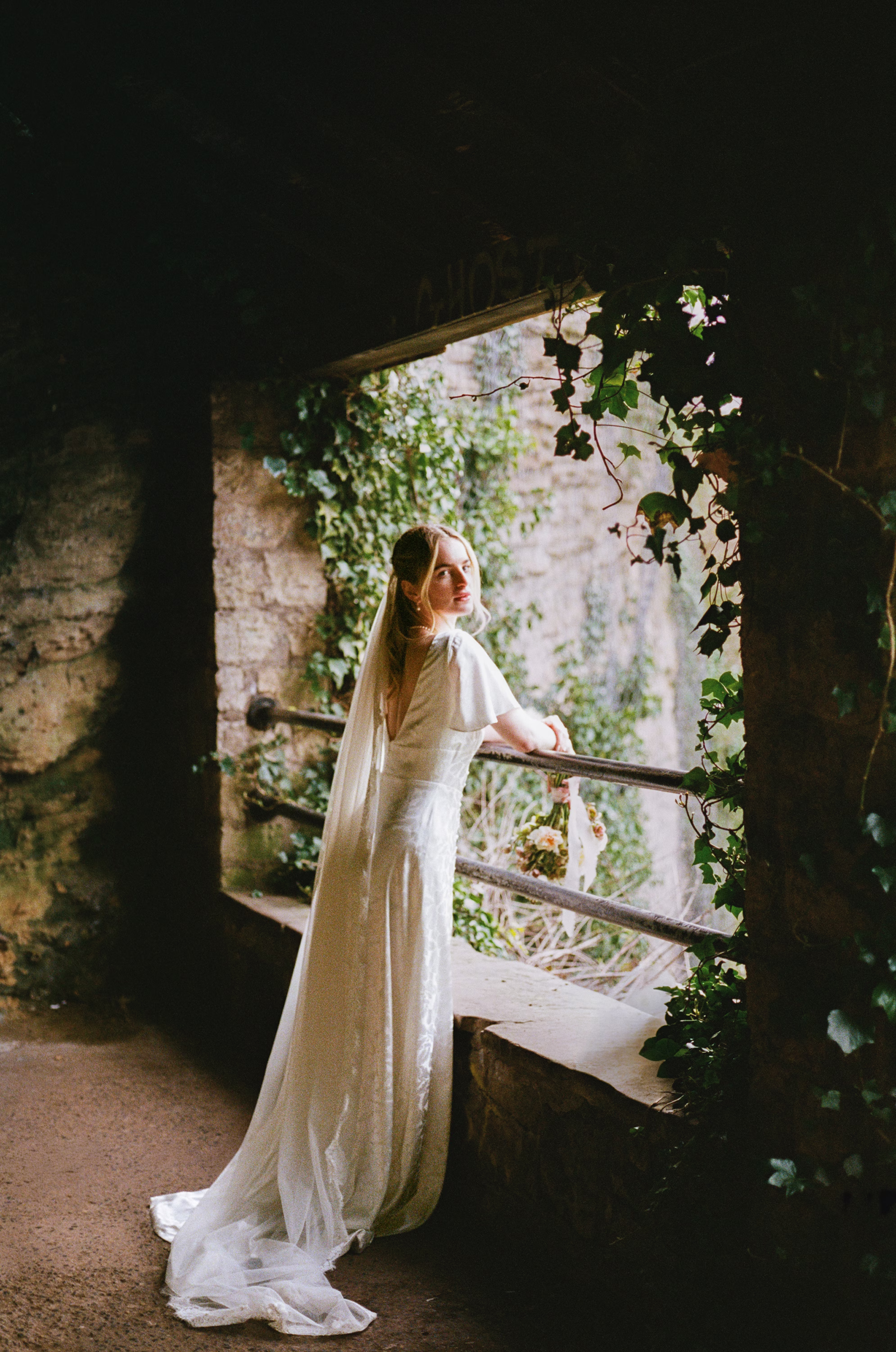 Bride in a flowing gown and long veil looking over her shoulder from an ivy-framed stone opening