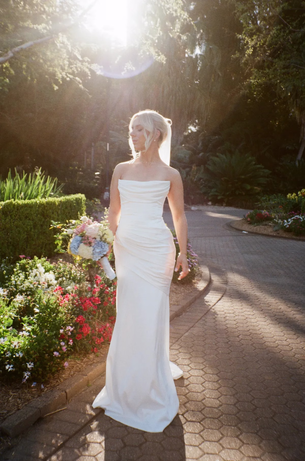 bride walking sunlit garden path the light photo and film - The Light Photo & Film: 35mm Wedding Photography and Super 8 for Creative Couples Bride in a fitted white dress carrying a bouquet along a garden path in late-afternoon sunlight.