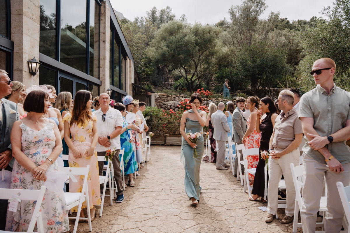 Emotional bridesmaid wiping a tear from her cheek as she walks down the outdoor aisle at a wedding in Mallorca, planned by Dybiec Wedding