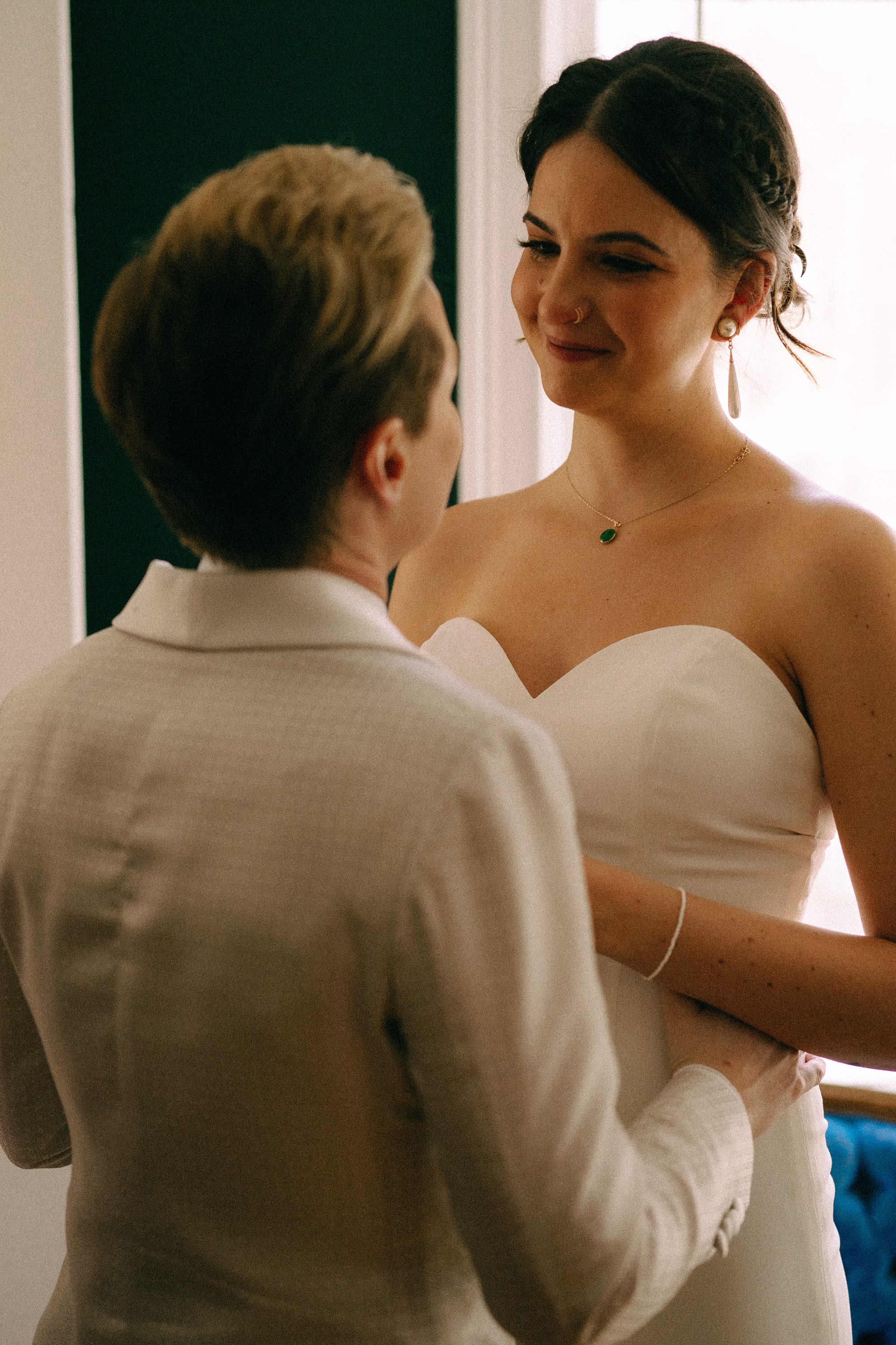 Bride wearing a sweetheart-neckline gown, drop earrings and a green pendant smiling at her partner beside a window