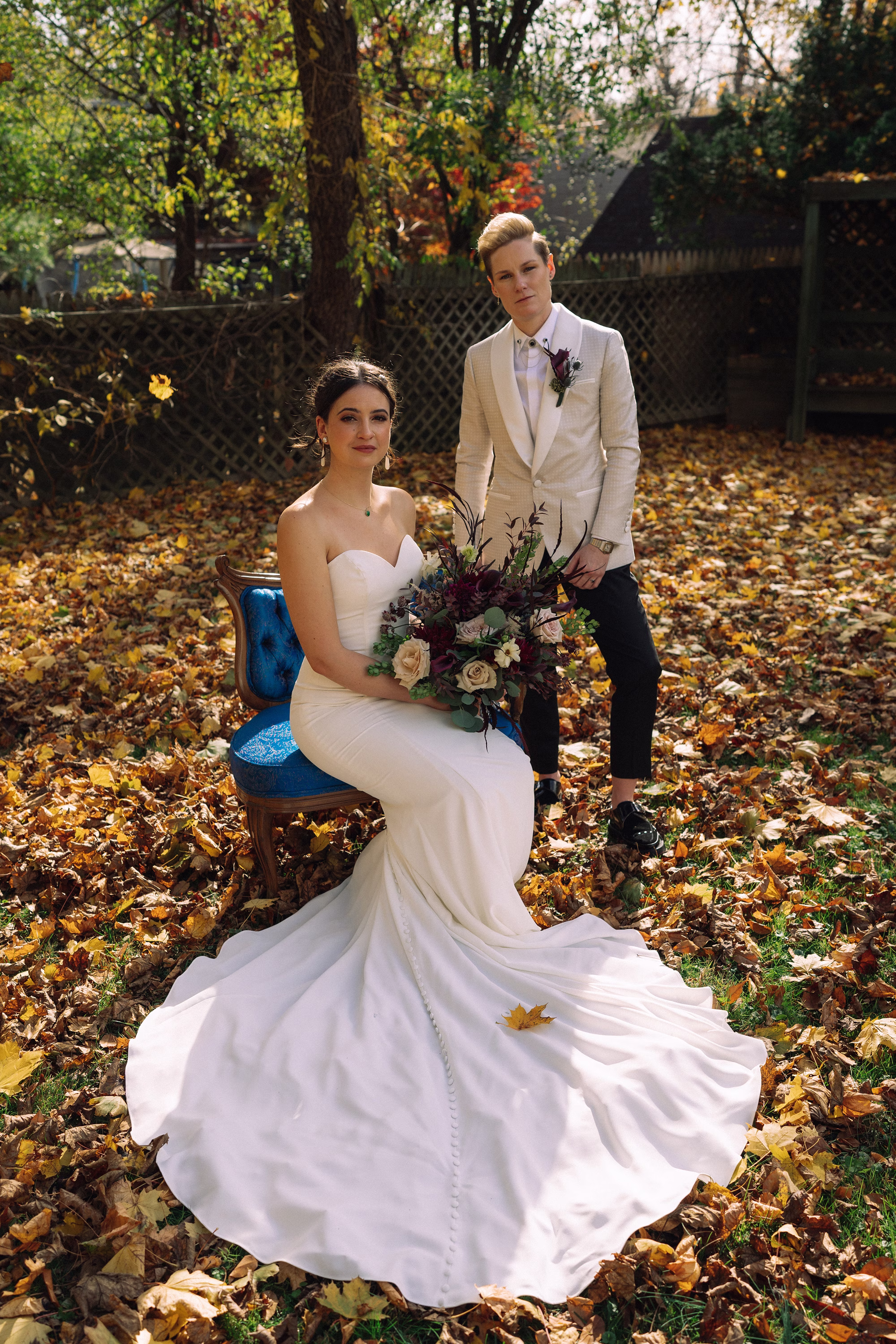 Bride seated on a blue velvet chair with her gown’s train spread over autumn leaves, her partner standing beside her in a white tuxedo jacket