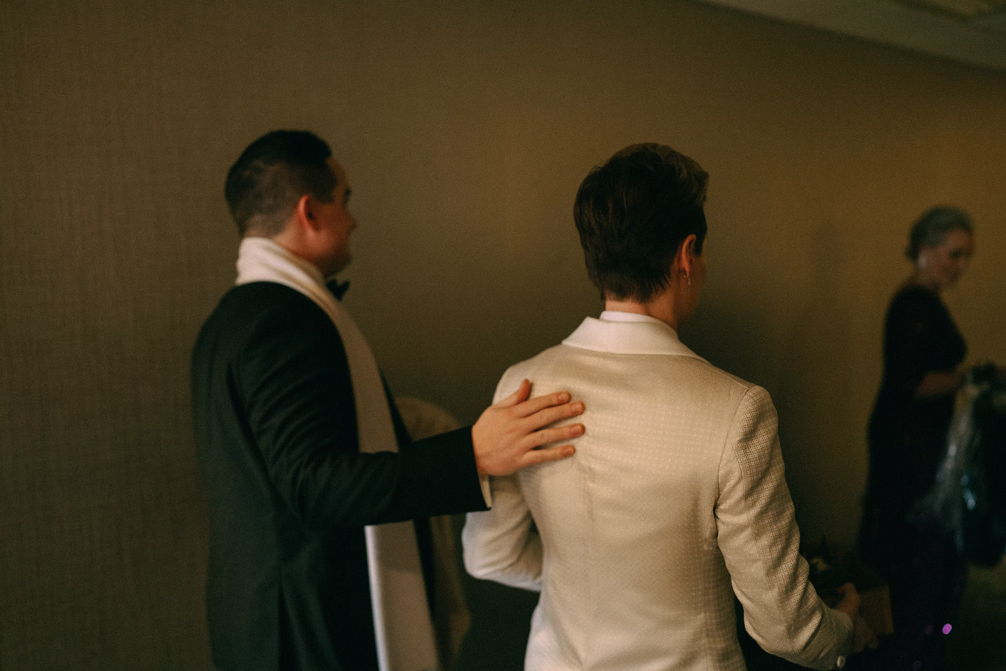 Back view of a bride’s textured white tuxedo jacket as a wedding party member rests a hand on her shoulder