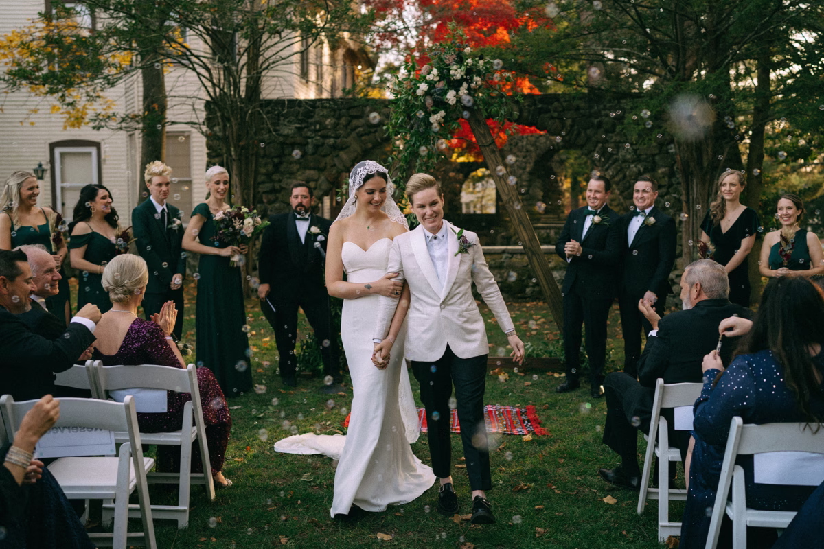 LGBTQ Newlyweds walking down the grassy aisle as wedding guests applaud and blow bubbles