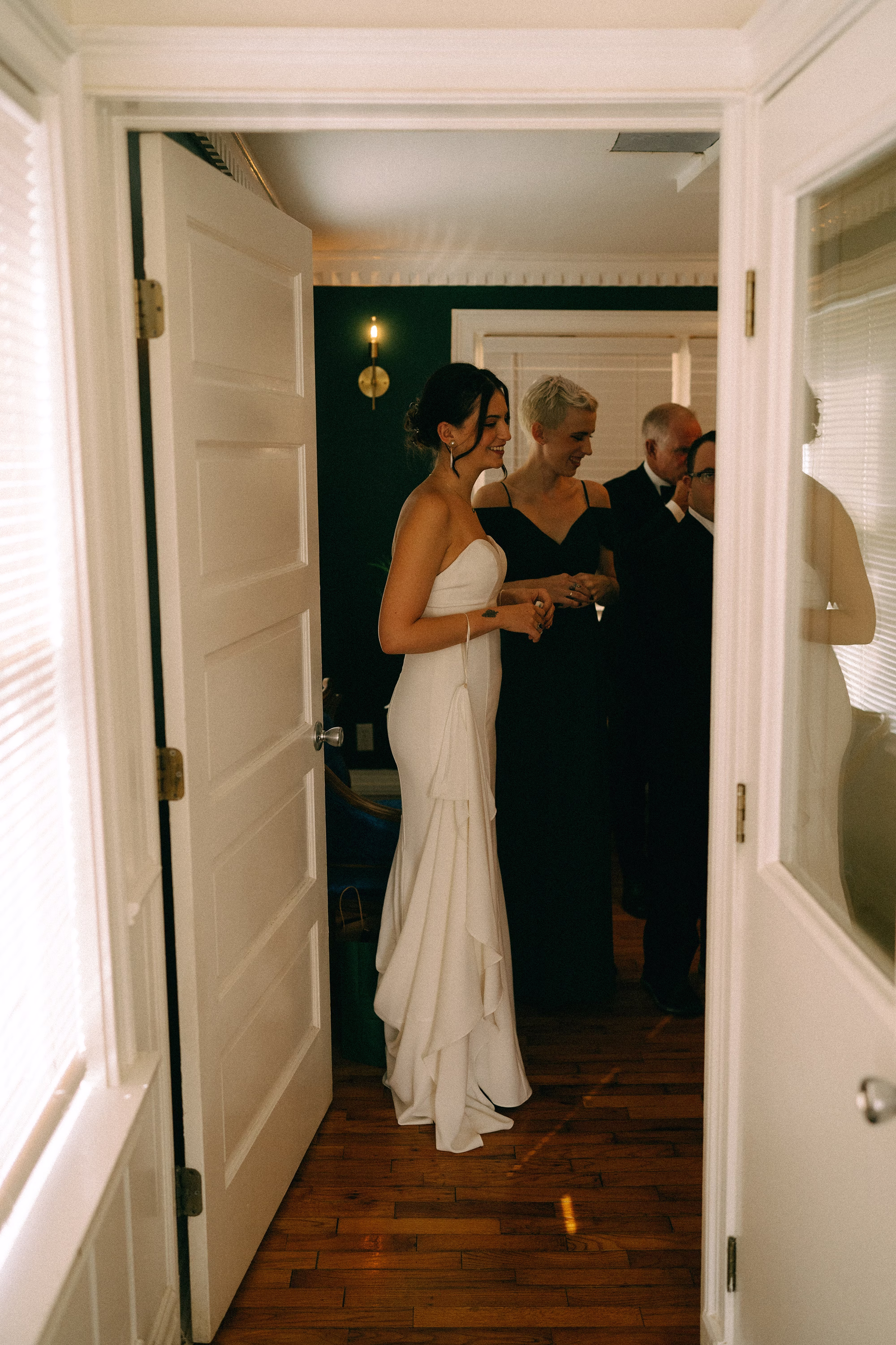 Bride in a fitted strapless wedding dress standing beside a bridesmaid in dark green, viewed through an open doorway