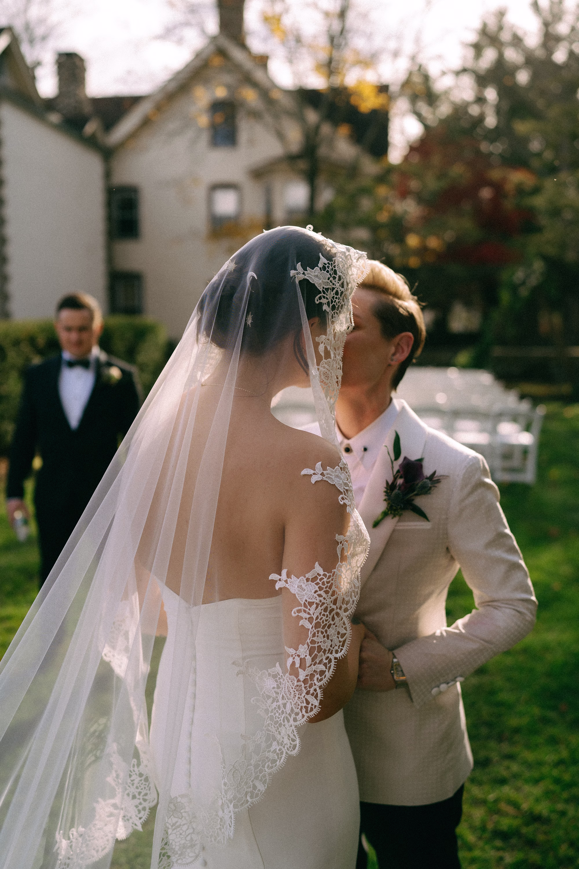 Bride in a floral lace-edged veil kissing her partner in a textured white tuxedo jacket outdoors