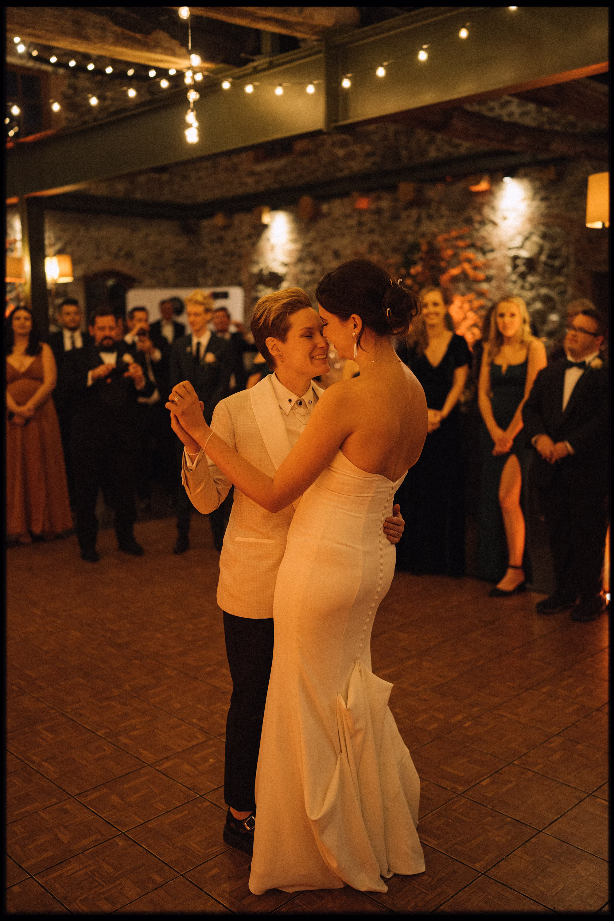 LGBTQ Couple sharing their first dance beneath festoon lights as wedding guests watch