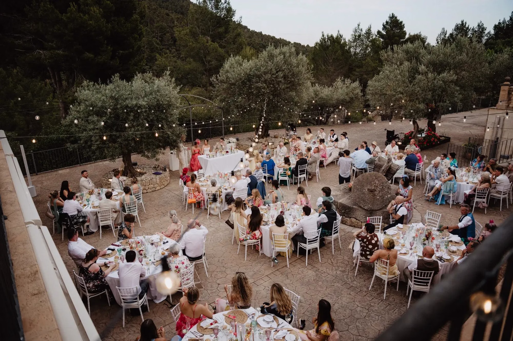 happiness mode photography 09 outdoor dinner - Getting Married in Mallorca From The UK: What You Need To Know Before You Book Anything View of a Mallorca wedding reception with festoon lights and guests seated at round tables, wedding planned by Dybiec Wedding